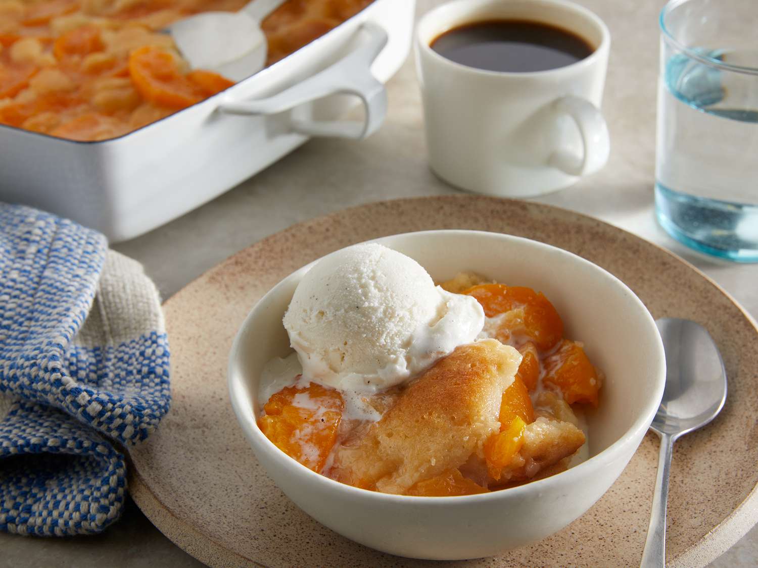 High view of peach cobbler, topped with melty vanilla ice cream and a cup of coffee in the background