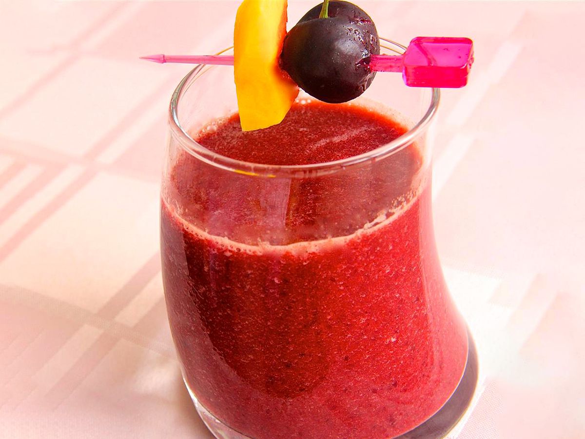 Close up view of a Mango Cherry Smoothie in a glass, garnished with a cherry and a piece of mango on a pink toothpick that's on top of the glass