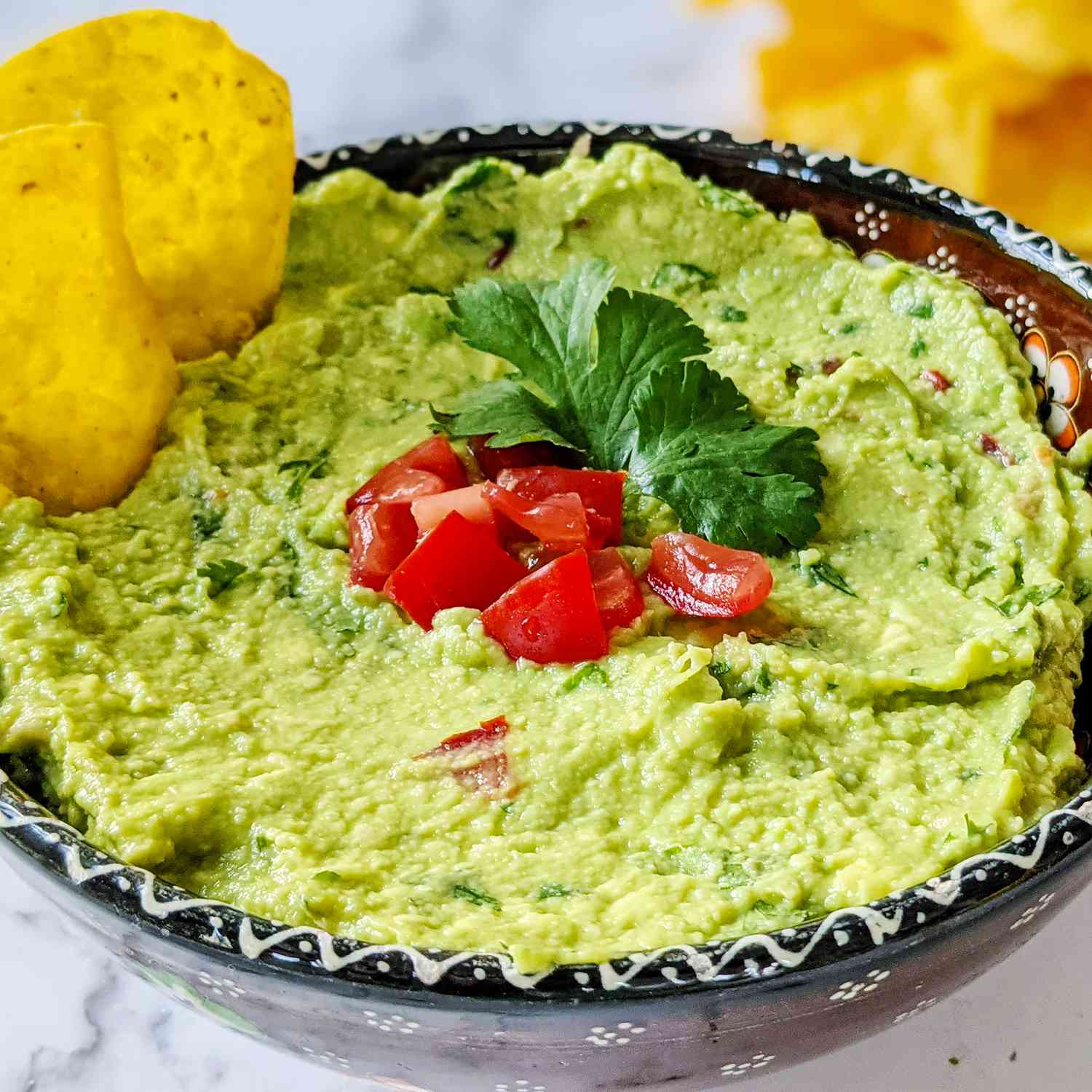 A low angle, close-up view of a bowl of fresh guacamole topped with diced tomatoes and parsley and served with tortilla chips.