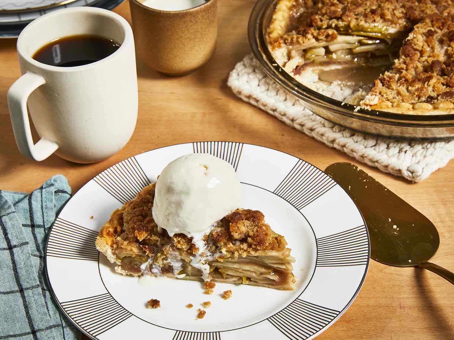 Close up view of a slice of Apple Crumb Pie with ice cream on top on a plate, with a cup of coffee and pie in a pie plate in the background