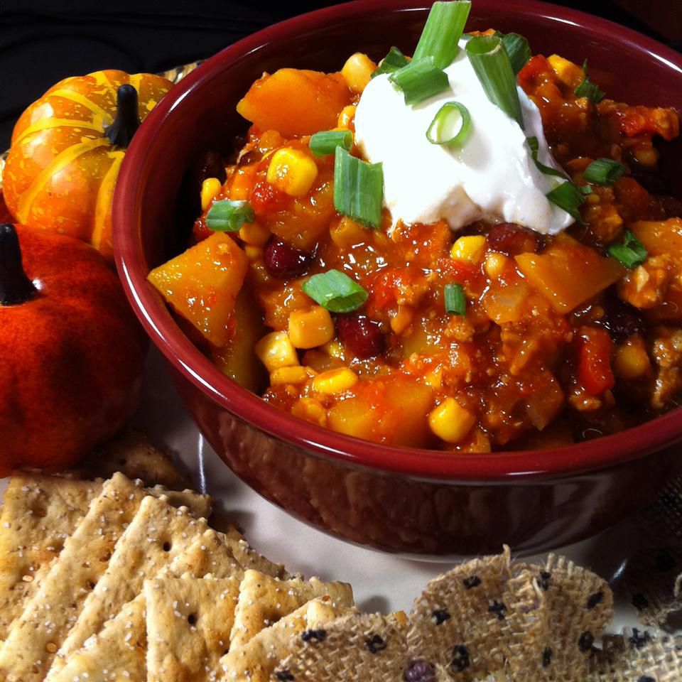 Close up view of Butternut Squash and Turkey Chili garnished with green onions and sour cream in a maroon bowl, next to crackers and mini pumpkins