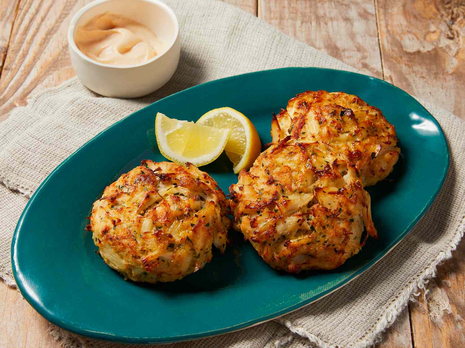 A high angle view looking at three large golden-brown crab cakes on a teal platter served with lemon wedges. 