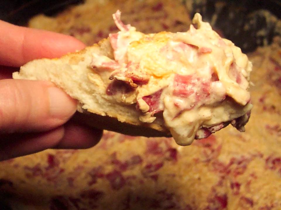 Close up view of Corned Beef and Swiss Dip on a piece of bread held by a hand, and dip in the background in a bowl
