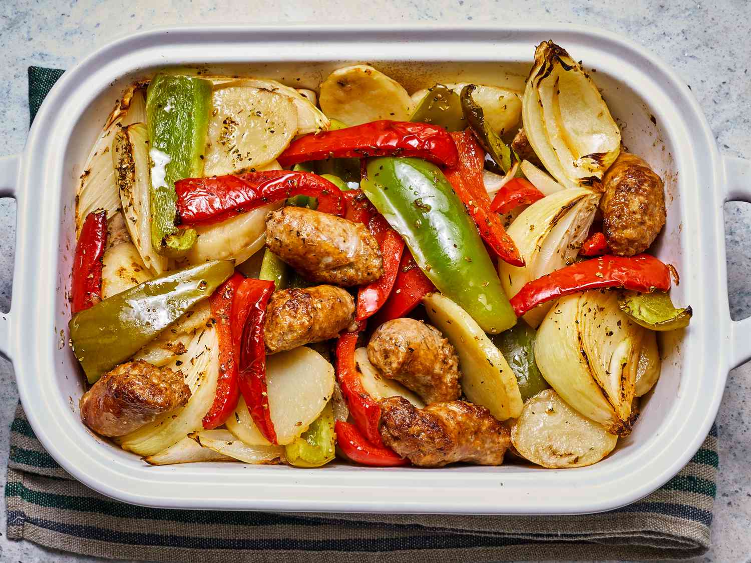 Overhead view of baked sausage, peppers, onions, and potatoes in a white baking dish.