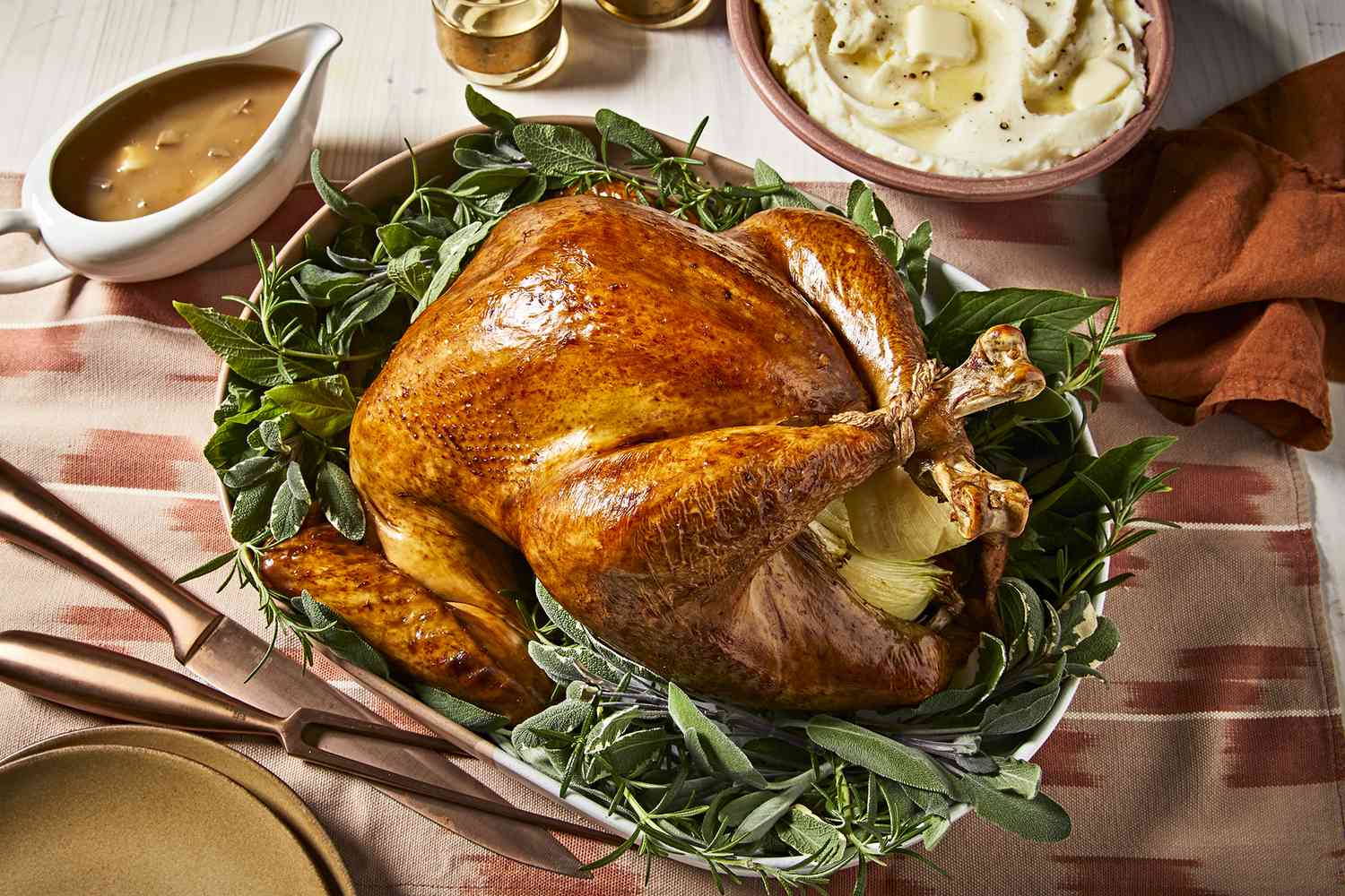 Overhead view of a perfectly roasted whole turkey served on a platter with lots of fresh herbs. The turkey is served alongside mashed potatoes and gravy