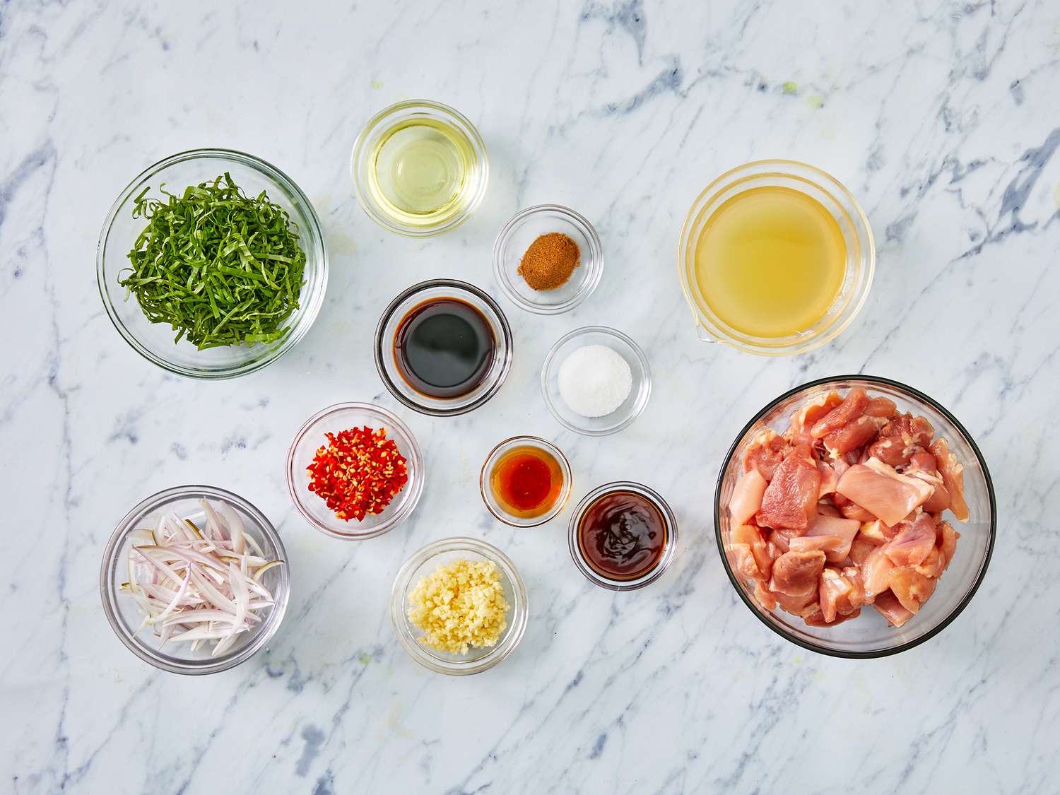 A top down view looking at ingredients for spicy basil chicken gathered in small glass bowls on a marble countertop. 