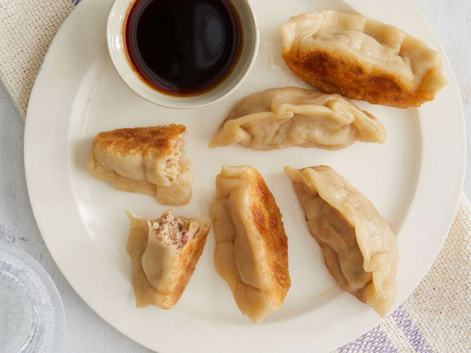 Looking down at a plate of golden brown potstickers with one cut open to see inside filling, with a side of dipping sauce