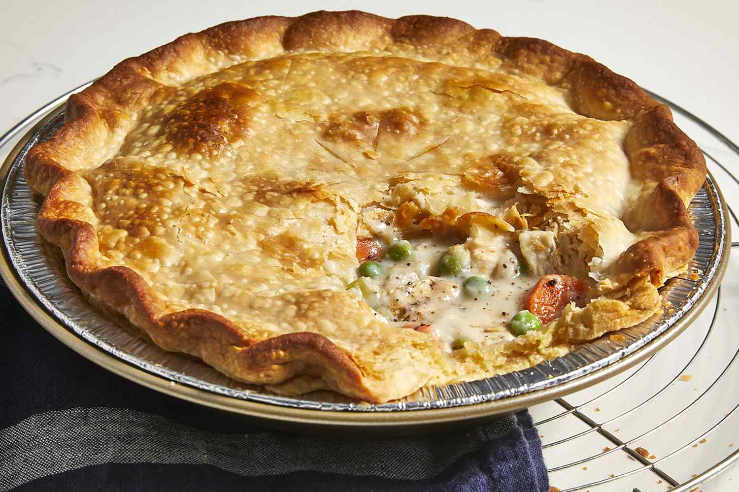 A low, close up view of a golden-brown chicken pot pie cooling on a rack, with a piece missing revealing the creamy hardy pie filling.