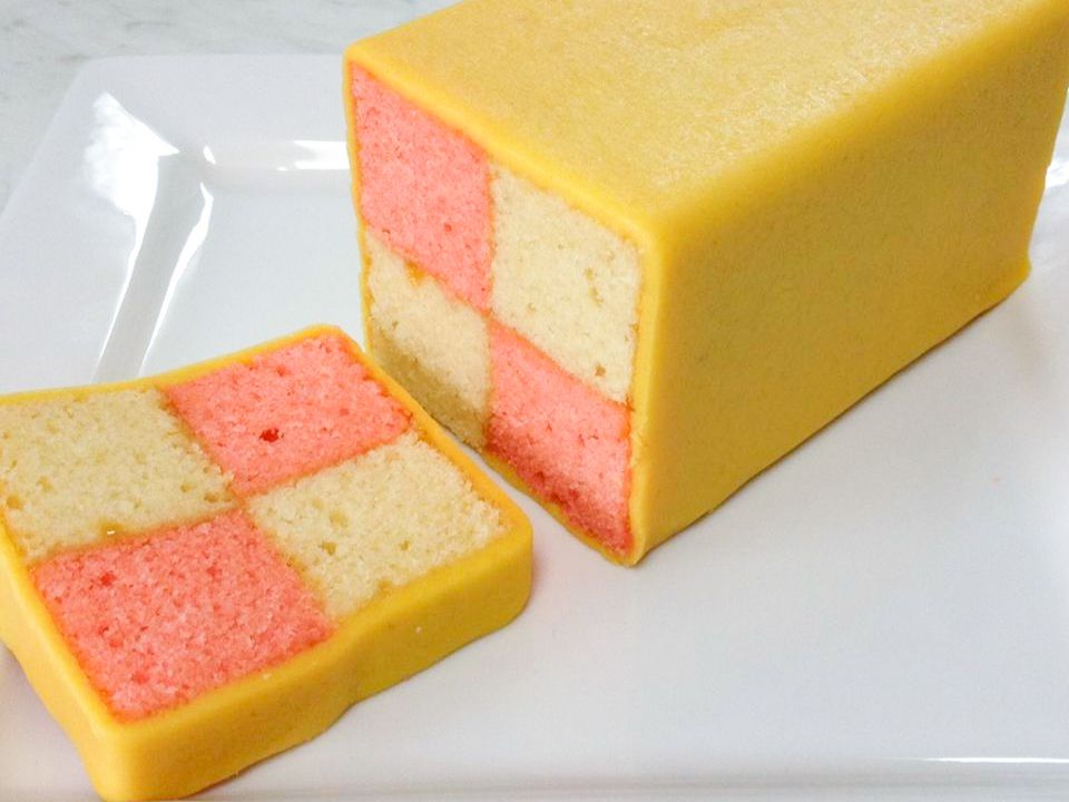Close up view of a slice of pink and white Battenburg Cake next to a loaf of Battenburg Cake that's yellow on the outside on a white plate