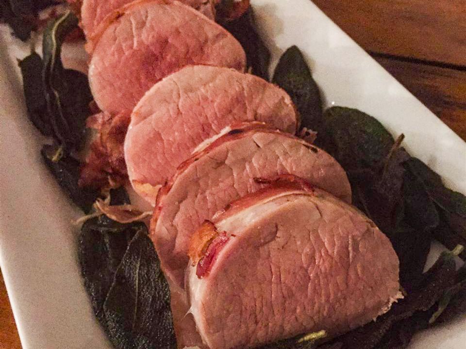 Close up view of a sliced Prosciutto-Wrapped Pork Tenderloin with Crispy Sage on a white platter