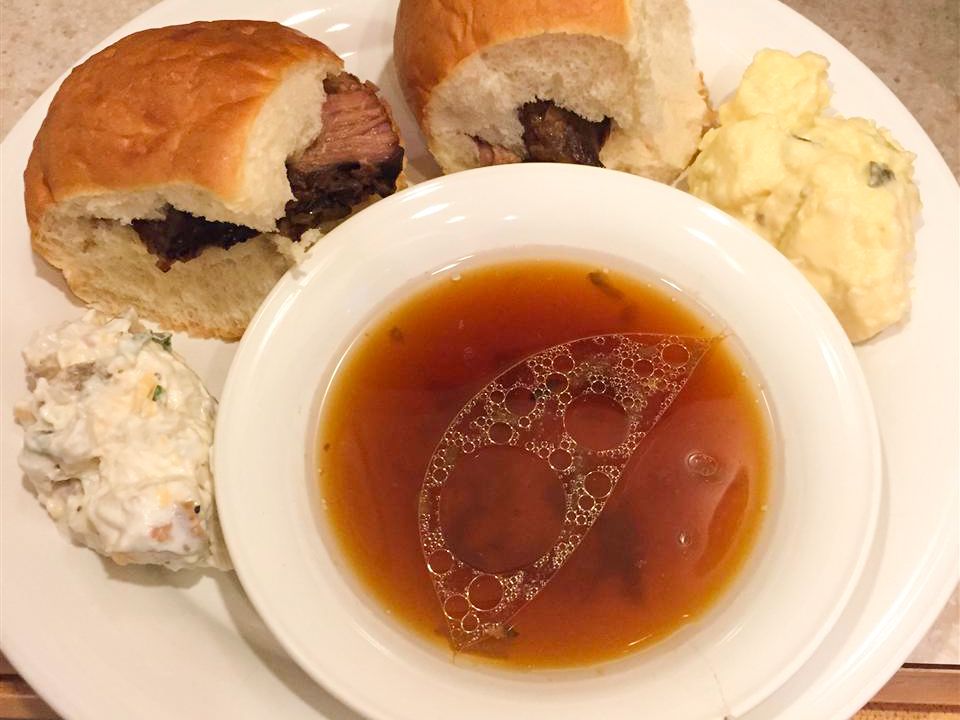 Close up view of French Dip in a white bowl, served with sandwiches and potato salad on a plate