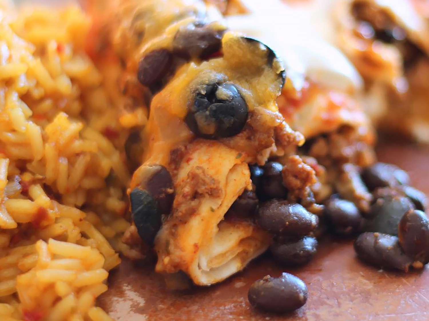 A super close up view of an enchilada with beef, black beans, and olives spilling out. The enchilada is served with rice. 