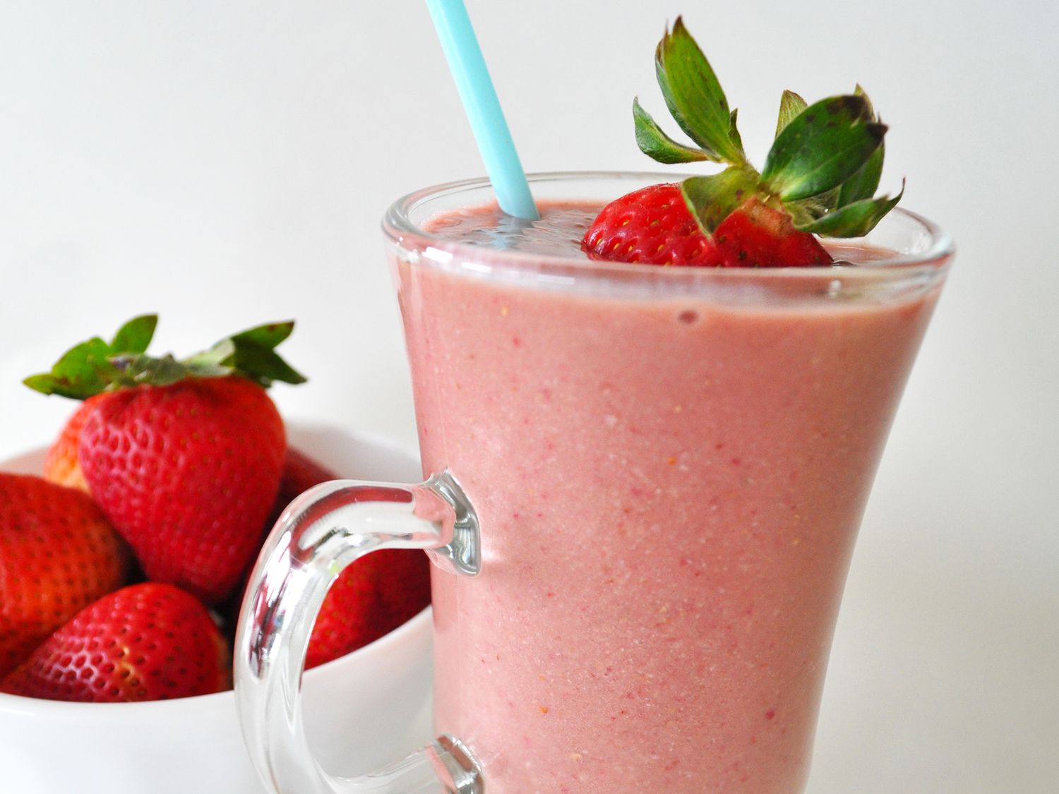 Close up view of a Strawberry Banana Protein Smoothie in a glass with a straw, garnished with a strawberry, and strawberries in a white bowl