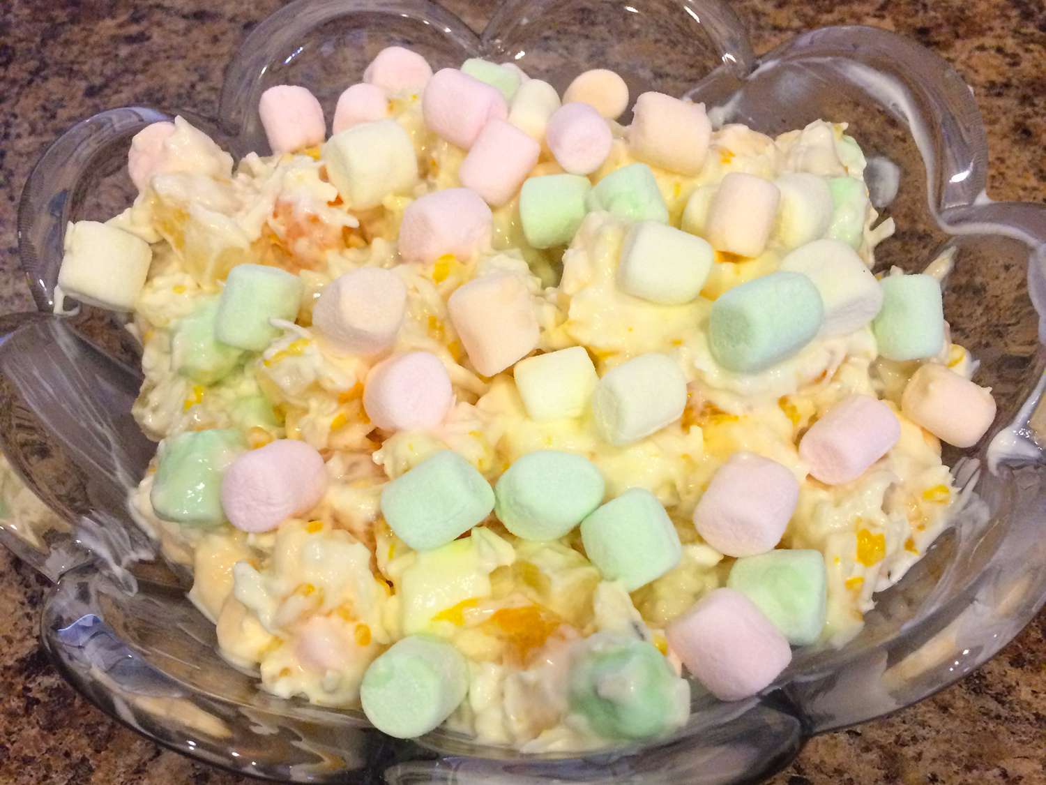 Close up view of a bowl with marshmallows, coconut pieces, sour cream, and mandarin pieces in a glass bowl