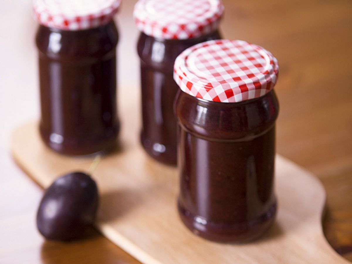 Close up view of Plum Butter in jars with red and white caps, next to a fresh plum on a wooden board