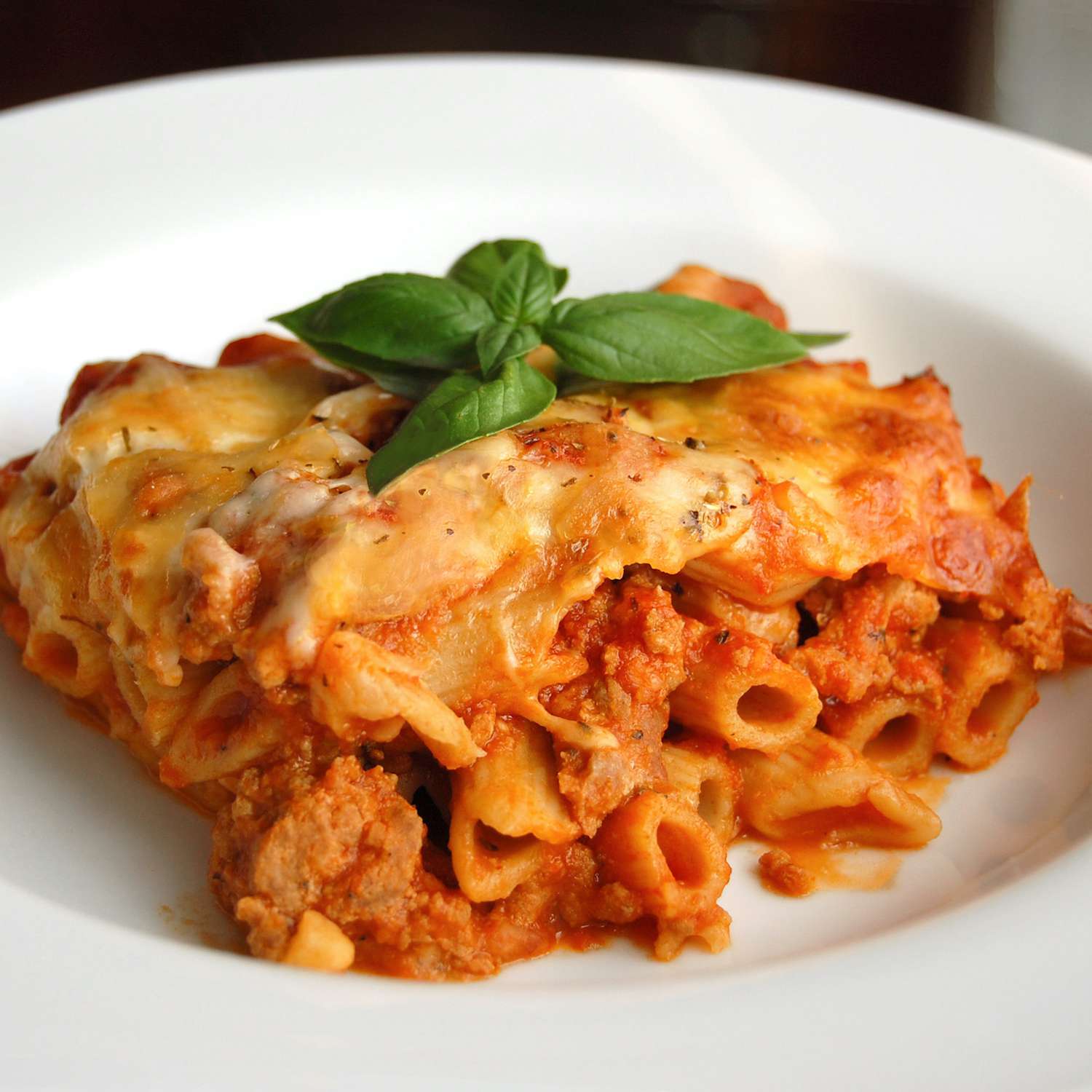 A serving of mozzarella-topped baked ziti in a white bowl, garnished with a basil sprig