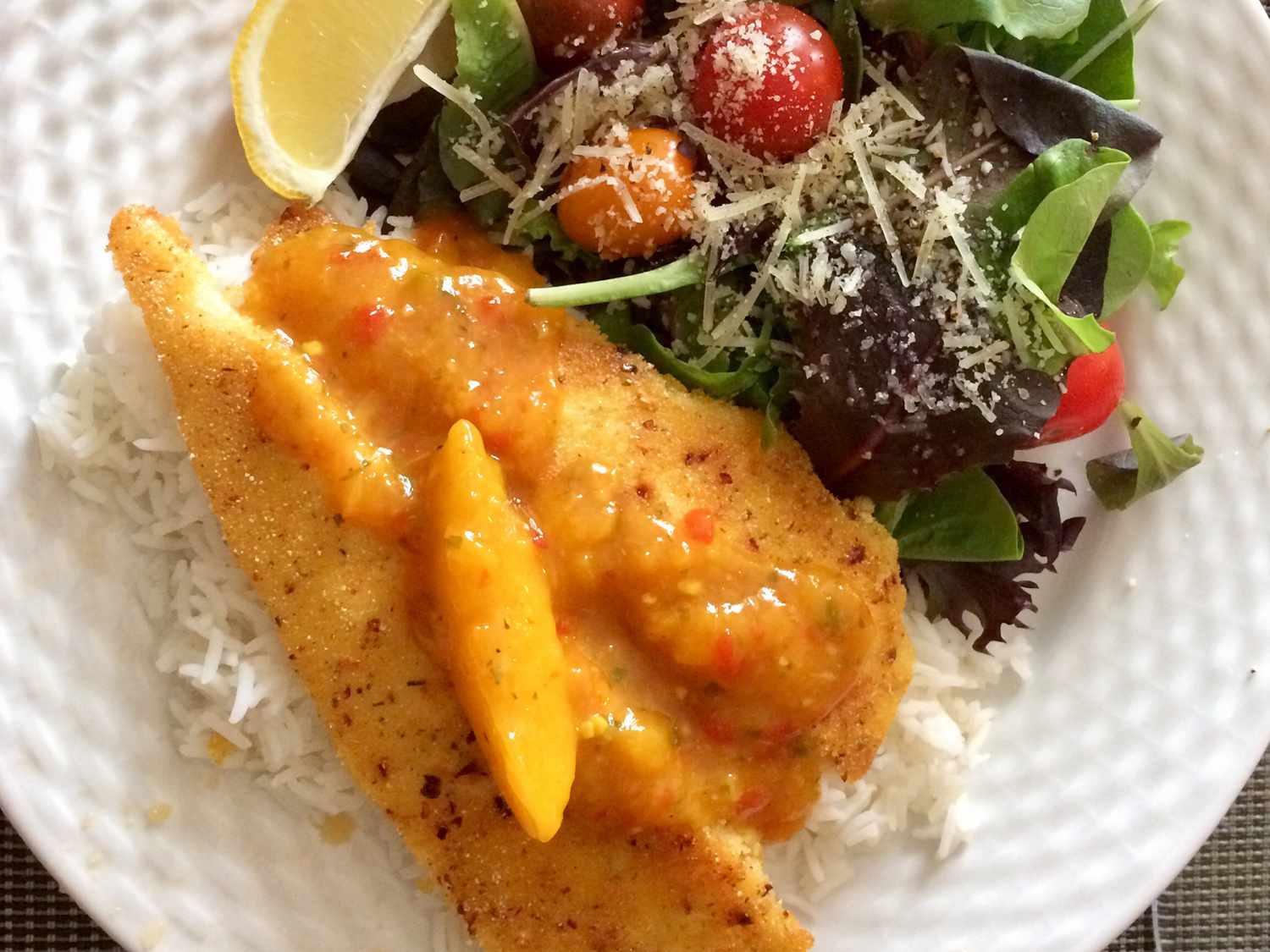 Close up view of a Pan-Fried Swai Fillet with peach sauce on white rice, served with salad topped with cheese and lemon wedge on a white plate