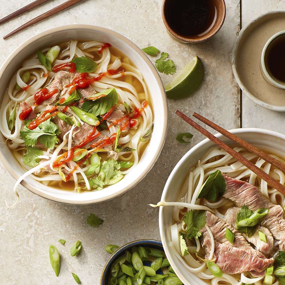 Close up view of Beef Pho garnished with fresh herbs and sriracha in bowls, with lime wedges, and chopsticks on a table