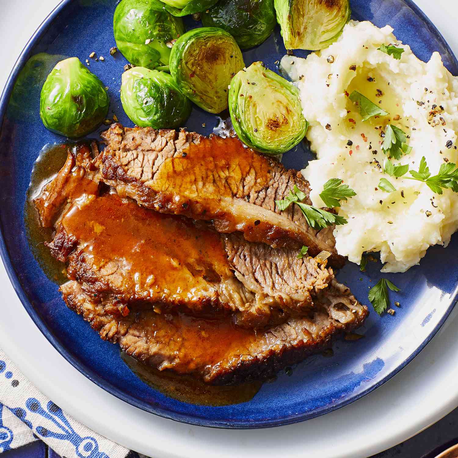 Close up view of sliced Jewish Style Sweet and Sour Brisket served with mashed potatoes garnished with fresh herbs and Brussels sprouts on a blue plate