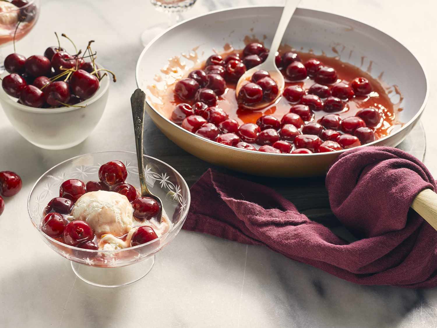 Close up view of Classic Cherries Jubilee in a serving bowl with ice cream, with cherries in a pan with a serving spoon, and fresh cherries in a bowl