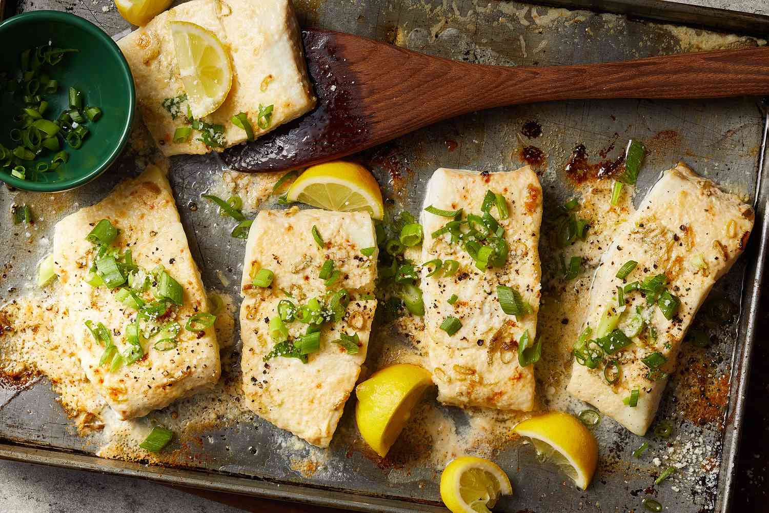 An overhead view of heavenly halibut filets on a baking sheet topped with sliced green onion and served with lemon wedges.