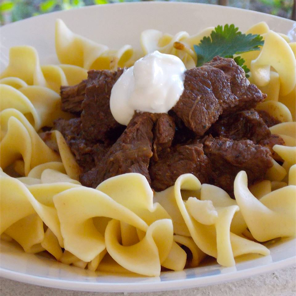 Close up view of Beef Goulash on top of pasta, topped with sour cream and fresh herbs in a white bowl