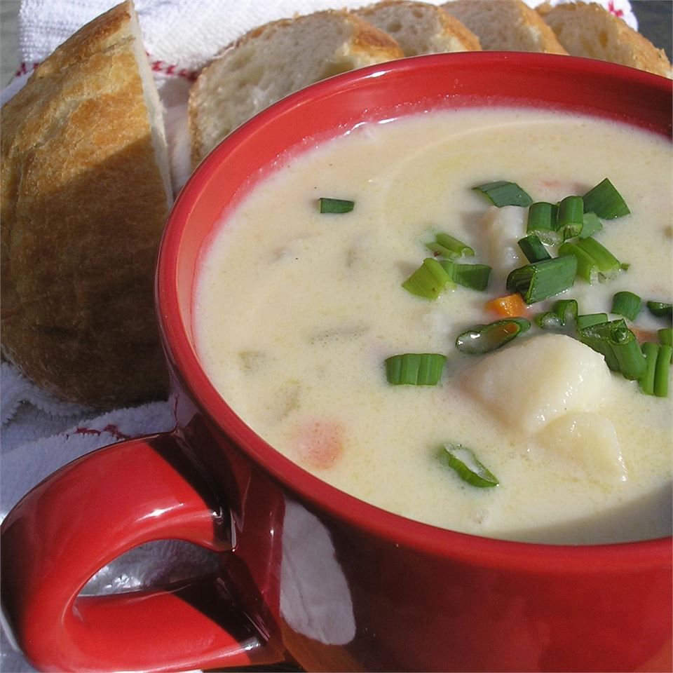 A red bowl of creamy soup with chunks of potato, garnished with sliced green onions with sliced bread in the background