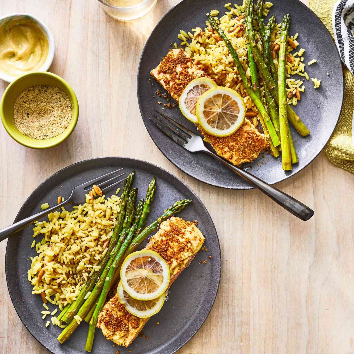 Breaded, baked salmon fillets topped with lemon slices, served alongside asparagus slices and rice pilaf on blue plates